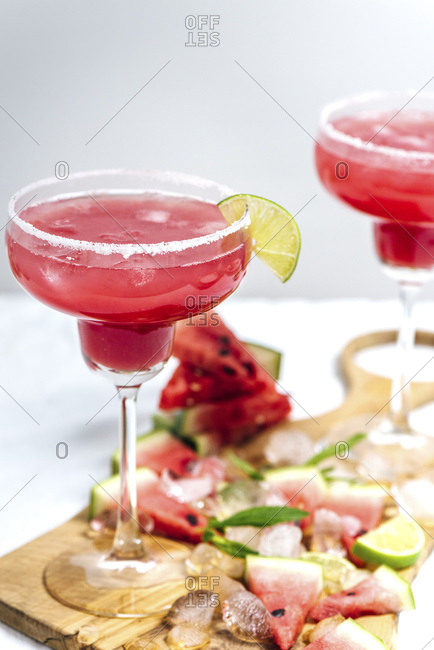 Watermelon margarita in margarita glasses standing on a wooden board photographed from front view. Ice cubes, watermelon slices, lime slices and mint leaves are on the board.