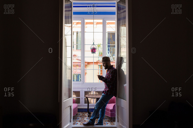 Side view of man in hat and checkered shirt standing in doorway of balcony and using phone.