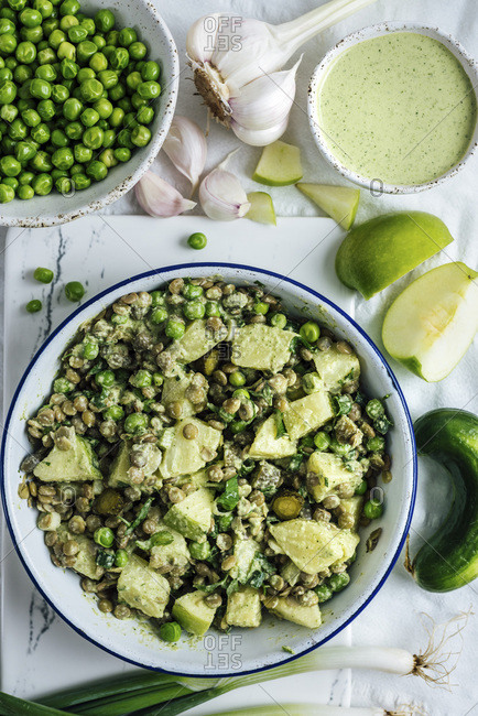 A bowl of potato salad with green lentils drizzled with tahini dressing on a white background photographed from top view Tahini dressing, peas, green apple slices, garlic and cucumber accompany