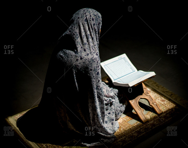 Muslim woman praying for god in dark room with Quran stock photo - OFFSET