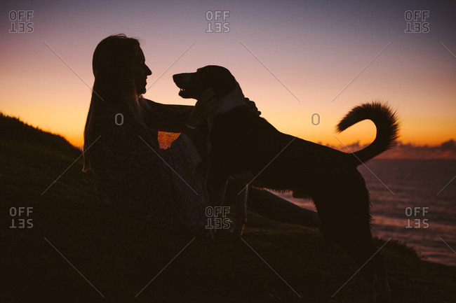 Side view of woman sitting with dog on hill against sky during sunset