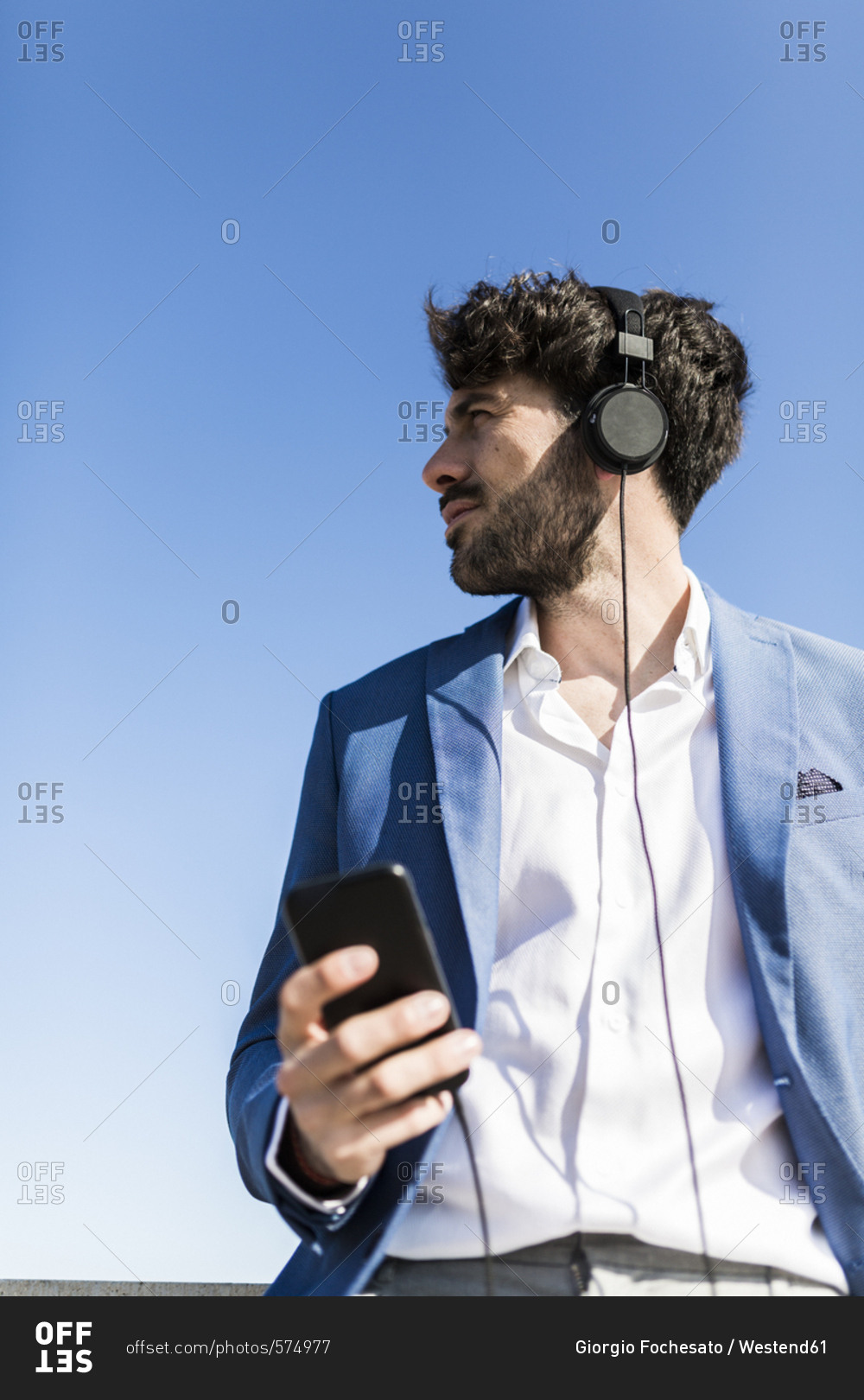 Young businessman with smartphone wearing headphones under blue sky