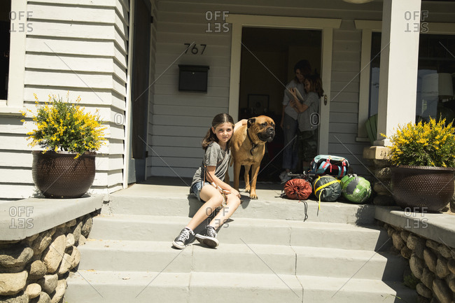 Girl sitting with dog and camping gear on steps with family standing at doorway in background