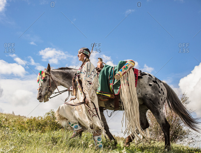 Native American girl wearing regalia walking with her horse on a hill