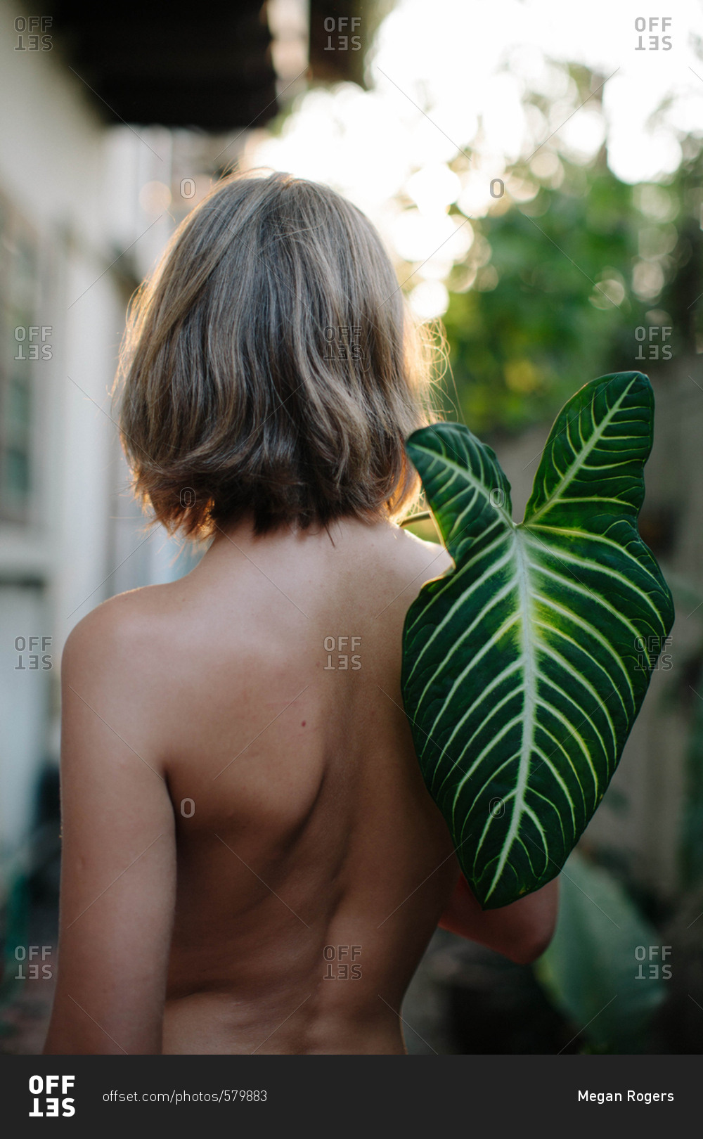 Young topless woman facing away from camera holding a large tropical leaf over her back