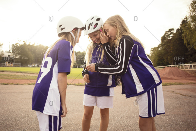 Happy female soccer players looking in mobile phone standing on footpath by soccer field