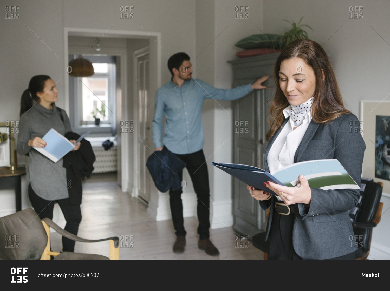 Smiling female real estate agent reading brochure while couple looking at cabinet at home