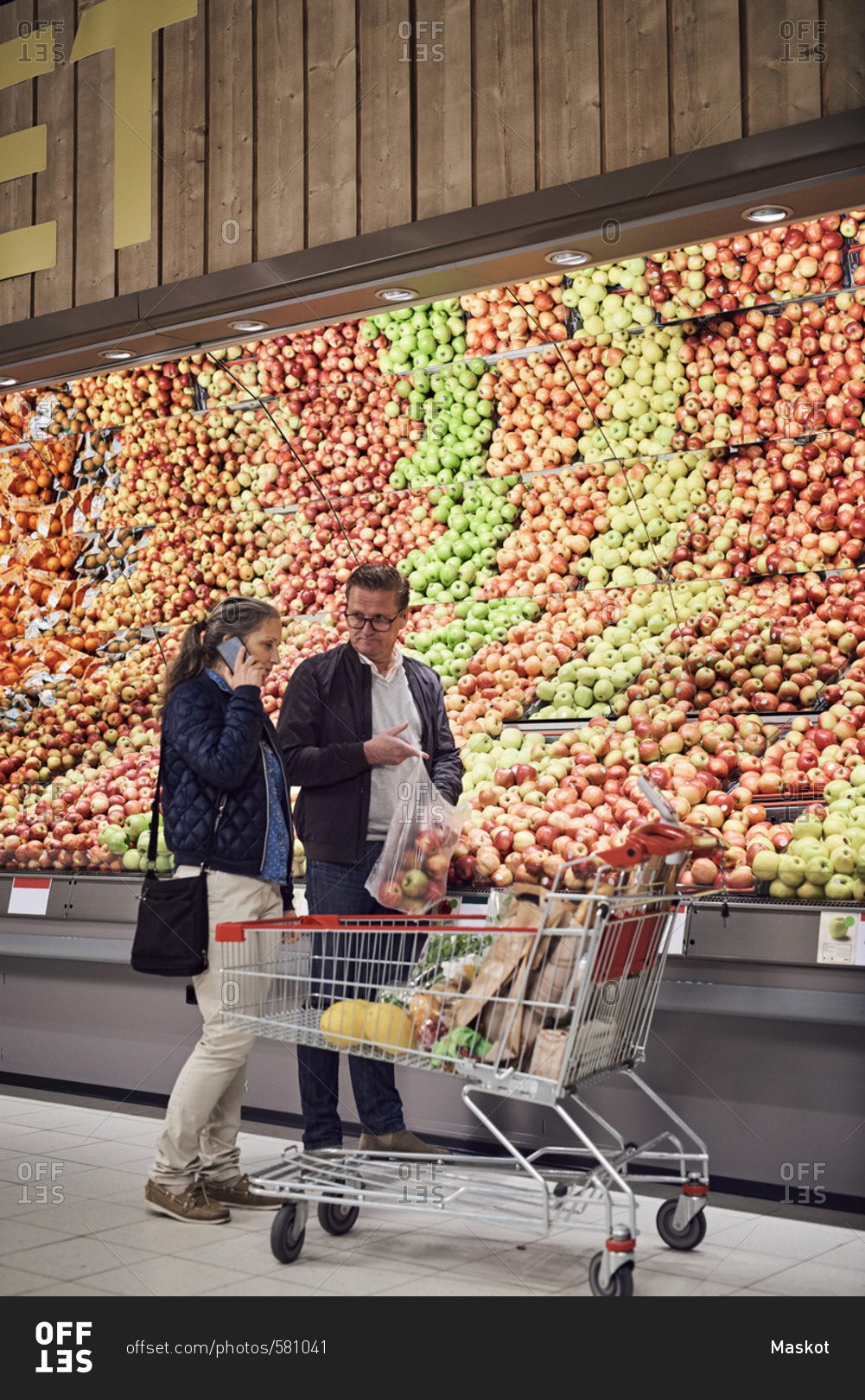 Man looking at woman talking on mobile phone while buying apples at supermarket