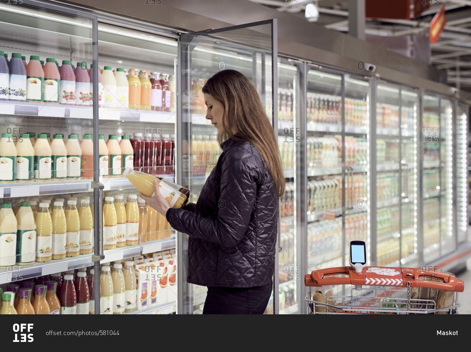Side view of woman holding juice bottles at refrigerated section in supermarket