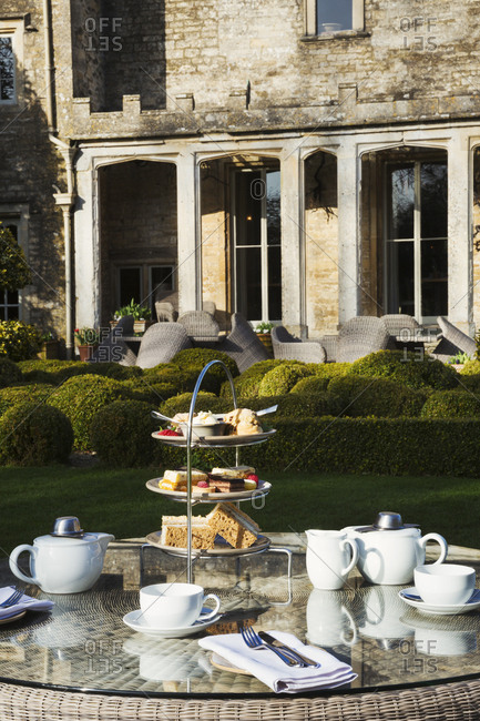 A table in a garden outside an old house, with cake stand and a selection of cakes and sandwiches set for a traditional afternoon tea