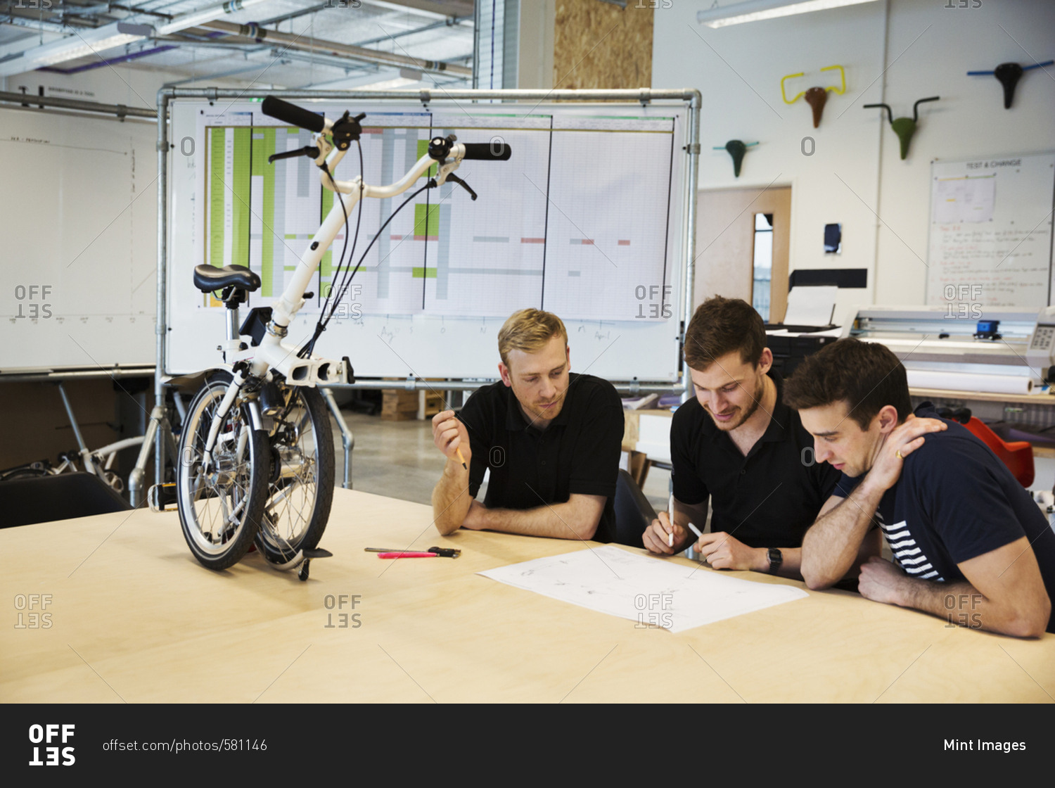Three men in a meeting at a bicycle factory, sitting at a table with a folding bicycle on the tabletop