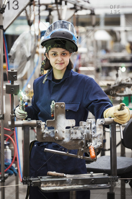 Female skilled factory worker, a young woman with a protective facemask lifted up, holding a welding tool, working on cycle frames