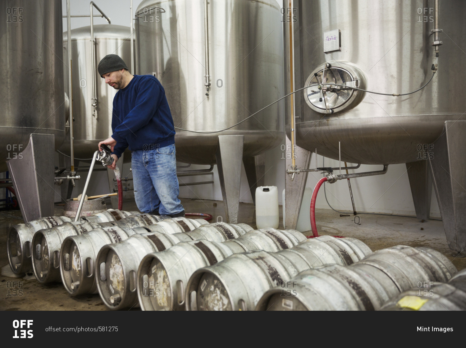 A man filling metal beer kegs from large fermentation tanks in a brewery
