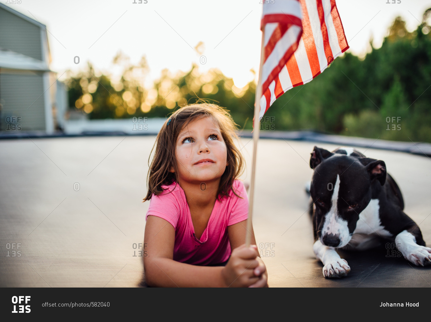 Girl looking up at American flag while lying on trampoline next to her pet dog