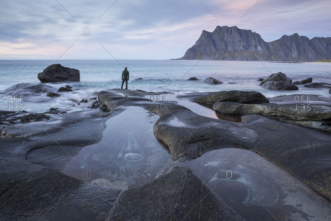 Man with beanie standing at the edge of a rocky beach and looking contemplatively towards a mountain in the distance