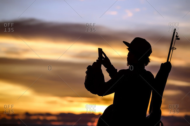 Silhouette of fisherman taking picture with smartphone at sunset