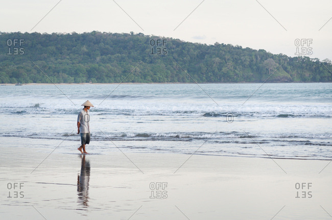 Pangandaran, Indonesia - May 22, 2011: A man wearing a woven conical rice hat strolls along the beach enjoying the calm ocean waves