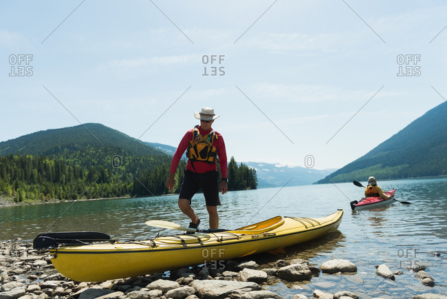 Man walking towards kayak with woman kayaking in background against sky
