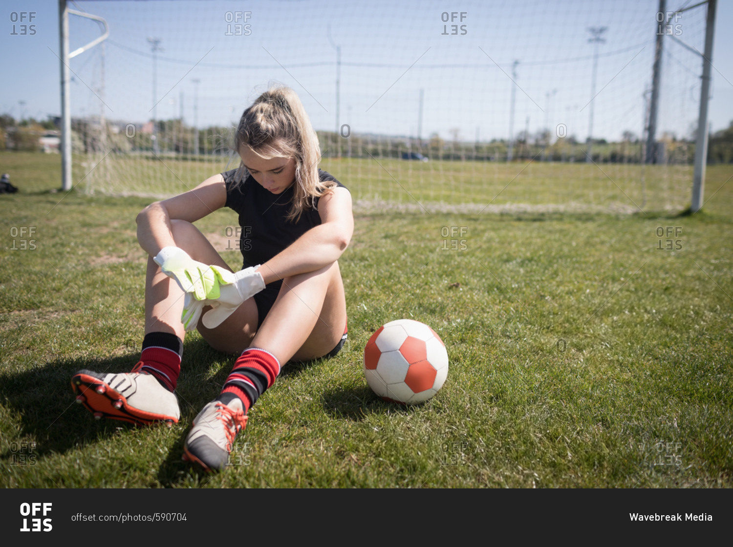 Full length of female soccer player resting on playing field during sunny day