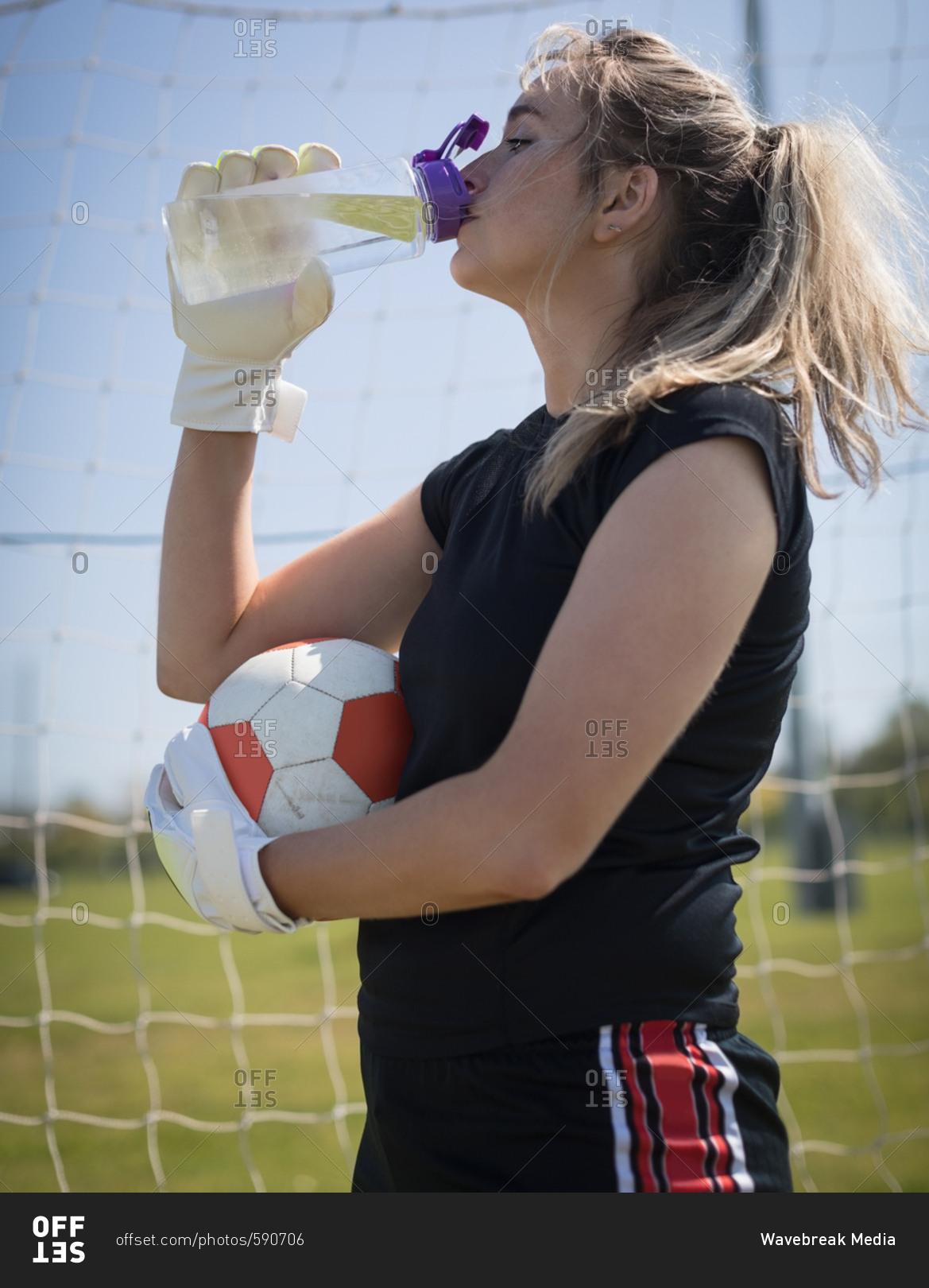 Thirsty young female soccer player drinking water while holding ball