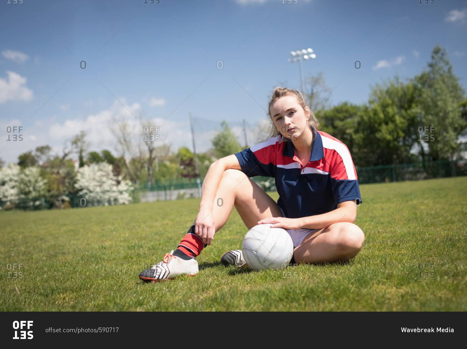 Portrait of young woman with soccer ball sitting on field against sky