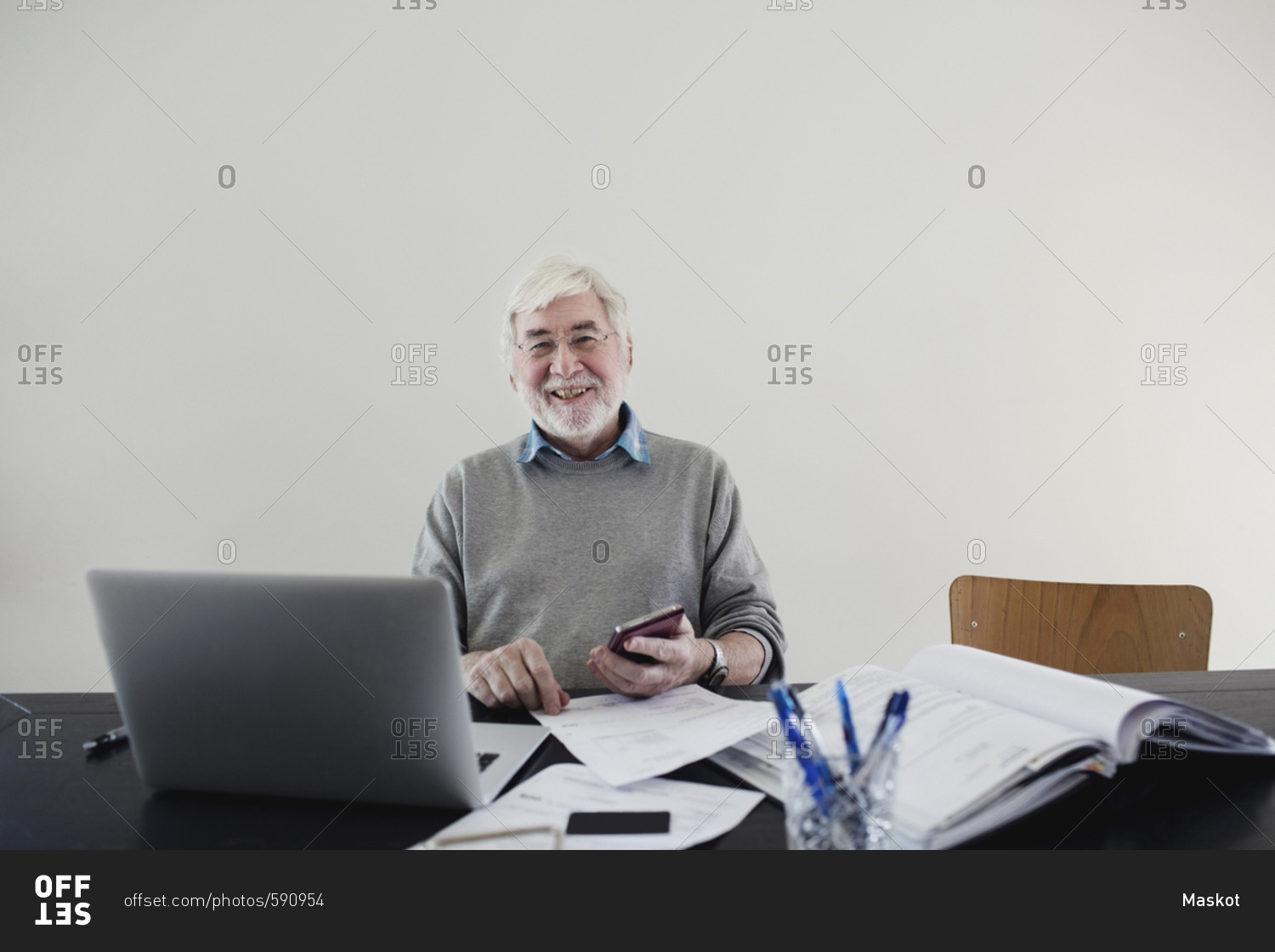 Portrait of smiling senior man with laptop and bills sitting against white wall