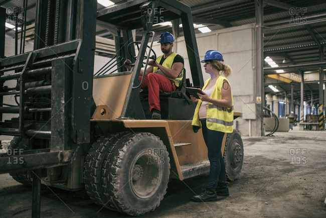 Wide angle image of workers in factory. Woman with tablet talking to forklift driver