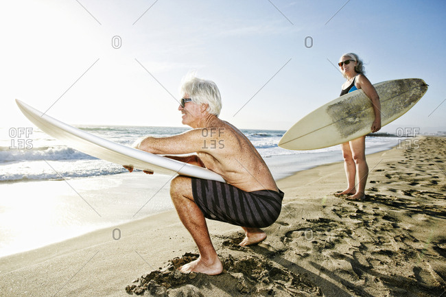 Older Caucasian couple standing on beach holding surfboards