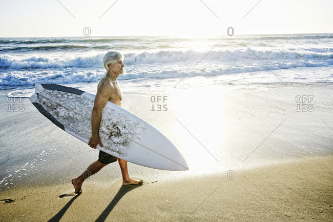 Older Caucasian man walking on beach carrying surfboard
