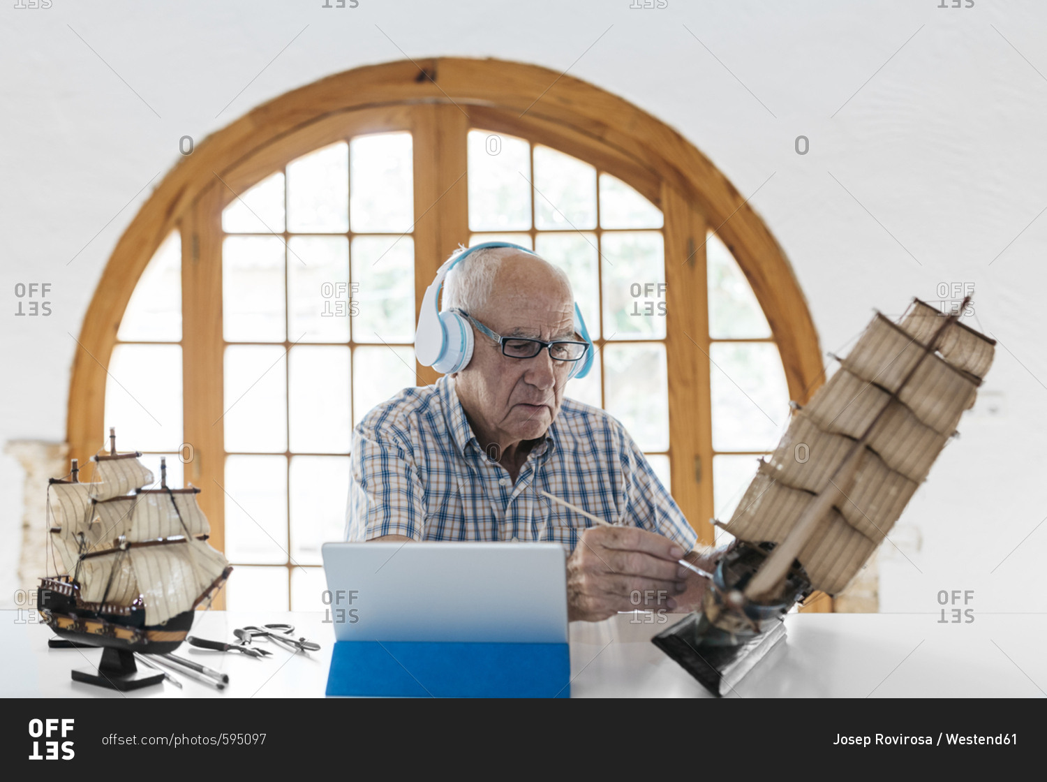 Senior man wearing headphones painting model ship on table with tablet