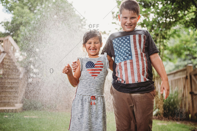 Brother and sister standing together in a sprinkler in a backyard