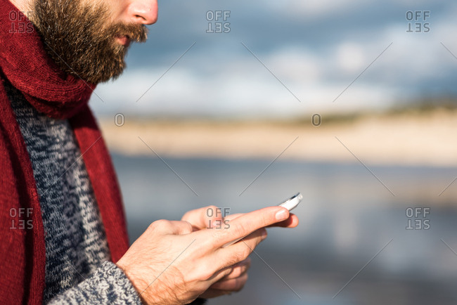 Crop faceless shot of bearded male in warm clothing using phone in nature.