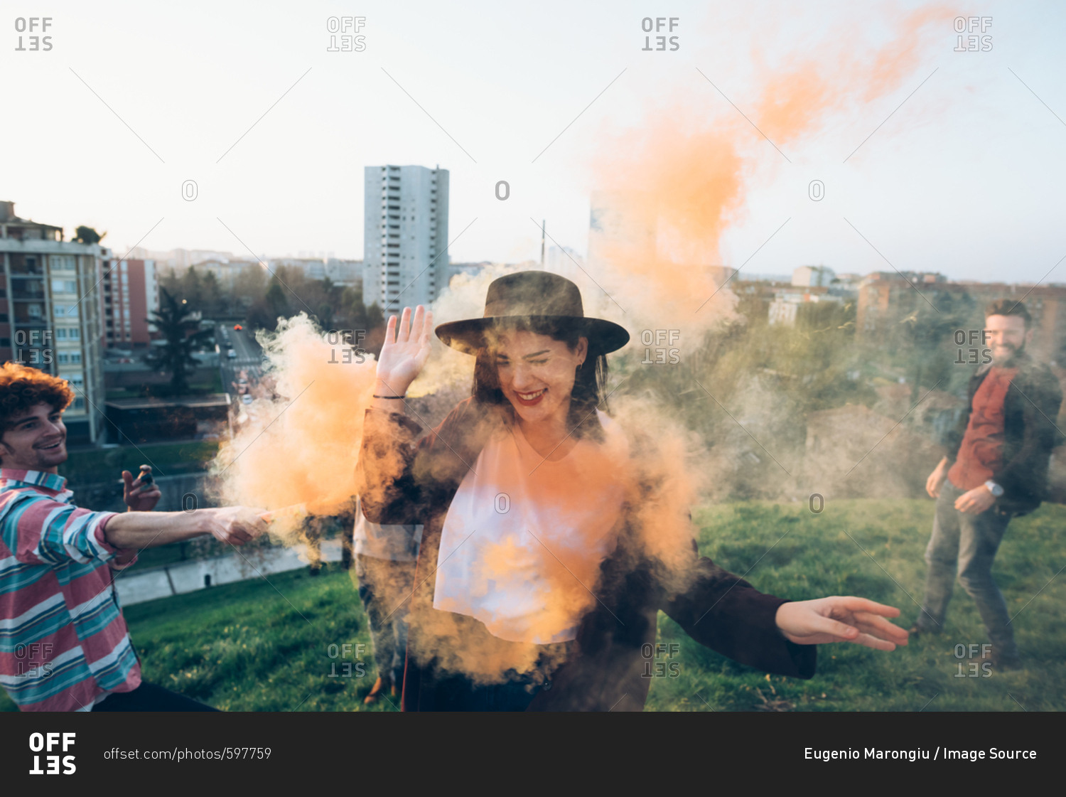 Group of friends on roof, holding colorful smoke flares, young woman walking through orange smoke