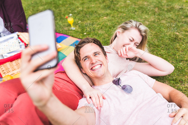 Group of friends relaxing on picnic blanket, in park, man taking selfie using smartphone