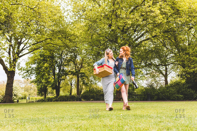 Two women walking across field, carrying picnic basket, blanket and bottle of champagne