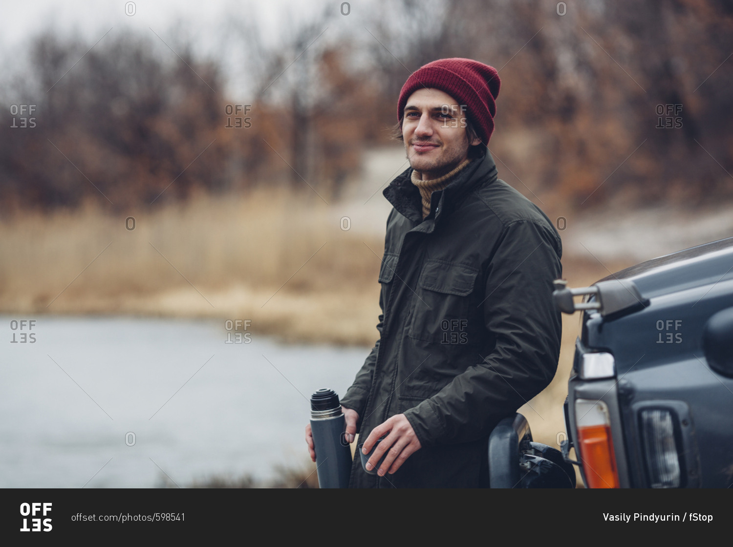 Smiling man holding insulated drink container while standing by sports utility vehicle at lakeshore