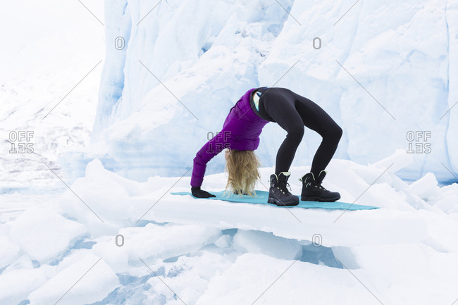 Young woman in winter clothes doing the upward bow or wheel yoga pose in front of Portage Glacier in the winter