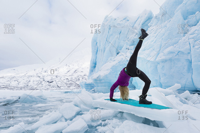 Young woman in winter clothes doing the one legged upward bow or wheel yoga pose in front of Portage Glacier in the winter