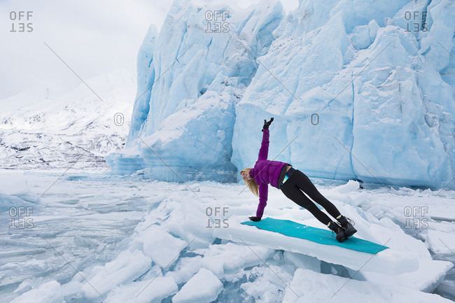 Woman in winter clothes doing the side plank yoga pose in front of Portage Glacier in the winter