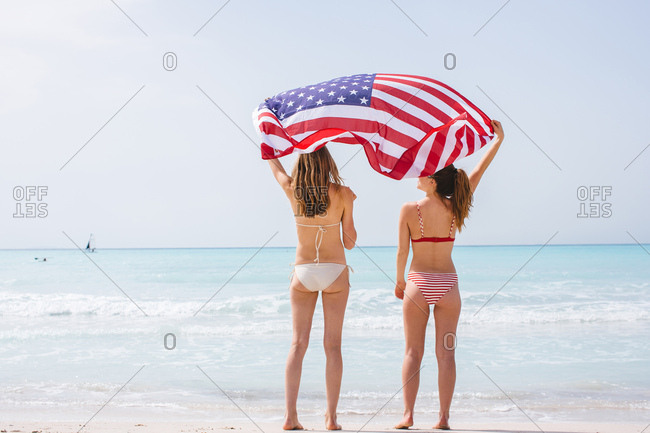 Rear view of two young female friends wearing bikinis on beach holding up American flag