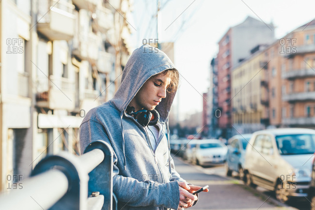 Man in urban area wearing hooded top and headphones looking down at smartphone