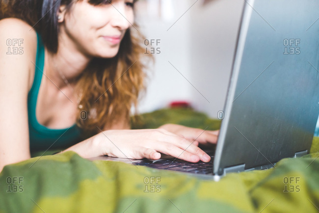 Young woman lying on front on bed using laptop computer smiling, differential focus