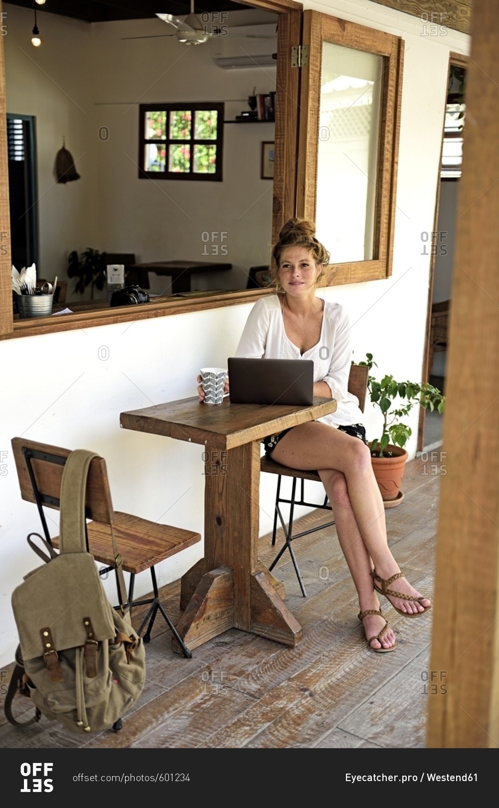 Portrait of young woman with laptop and mug of coffee sitting on veranda of a coffee shop