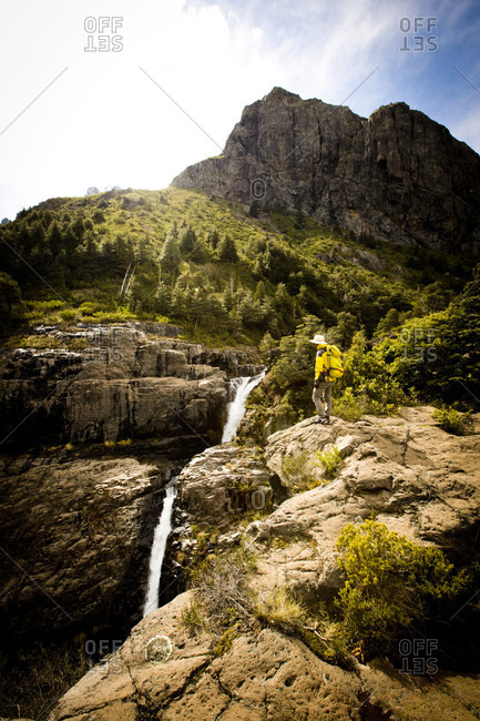 Climber wearing a vibrant yellow jacket and backpack, overlooks a waterfall flowing through the canyons. Lakes District, Chile