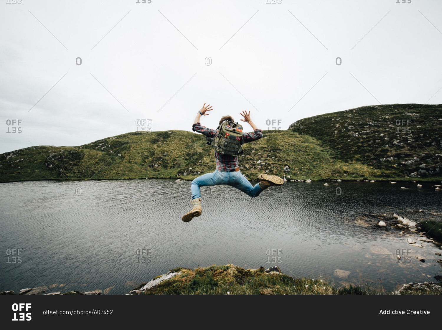 Back view of man with backpack jumping above ground on background of lake and mountains