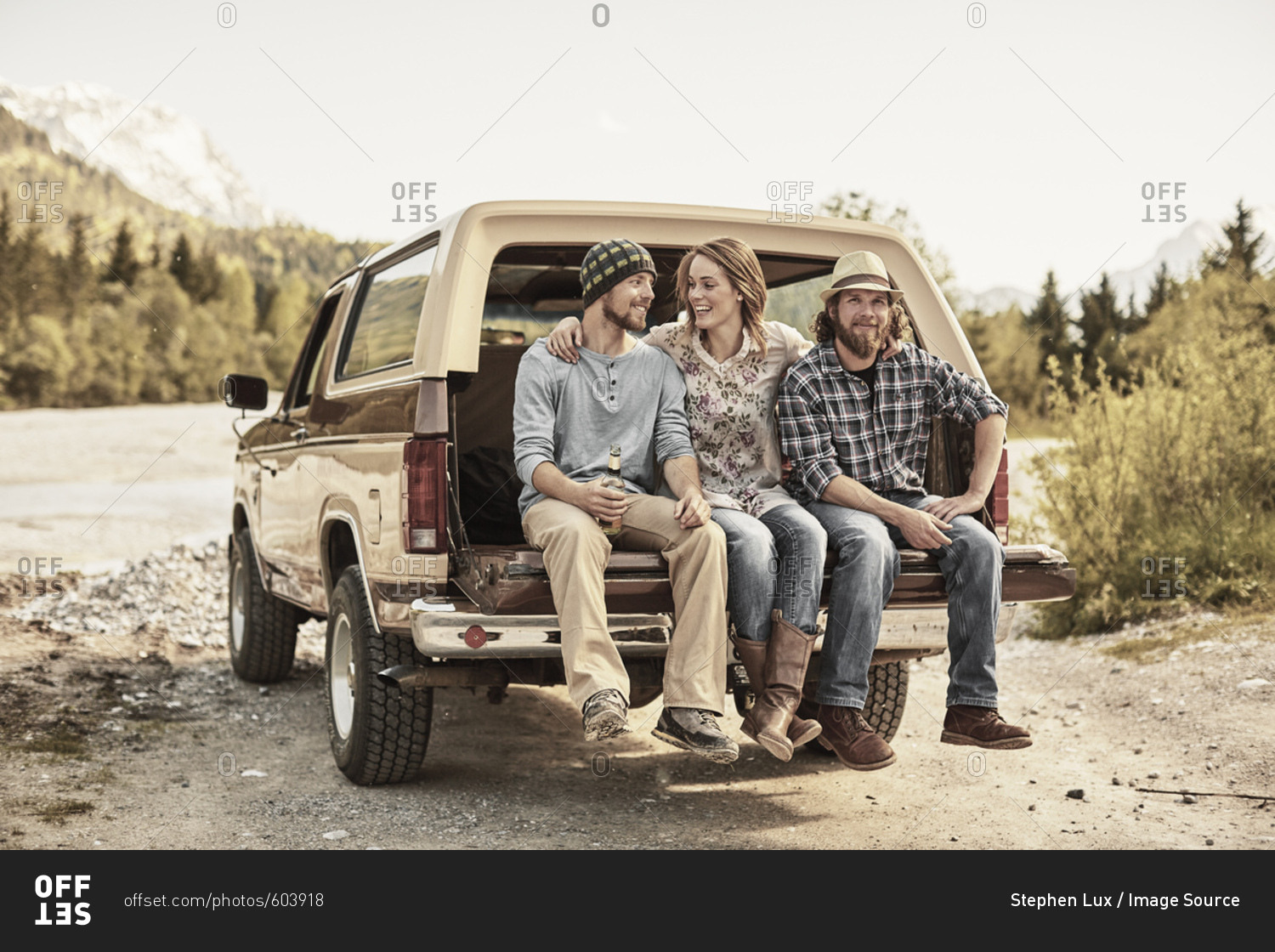 Three people sitting on back of pickup truck, arms around shoulders, smiling