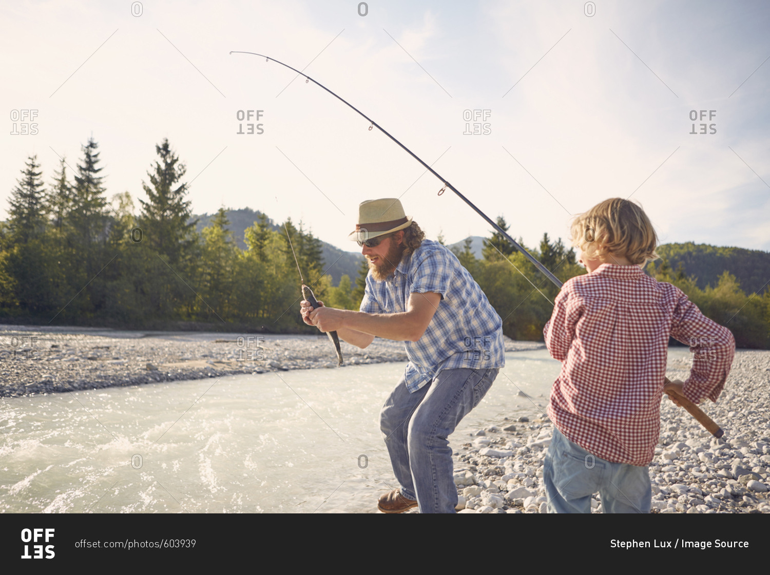 Mid adult man and boy next to river using fishing rod to catch fish