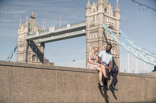 Young couple sitting on wall, smiling, Tower Bridge in background, London,  England, UK stock photo - OFFSET