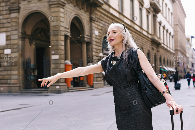 Mature woman with long grey hair with wheeled suitcase hailing a cab in Florence, Italy