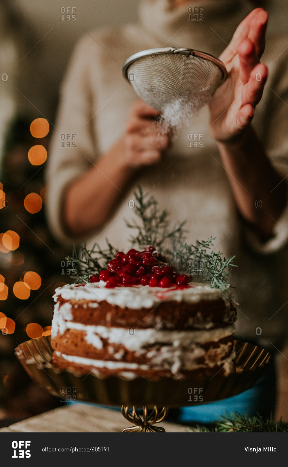 Woman decorating winter inspired naked cake with powdered sugar with Christmas tree in the background